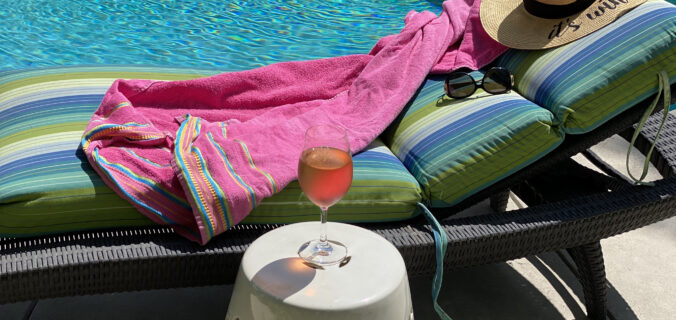 A glass of rosé next to a pool and lounge chair with sunglasses and straw hat